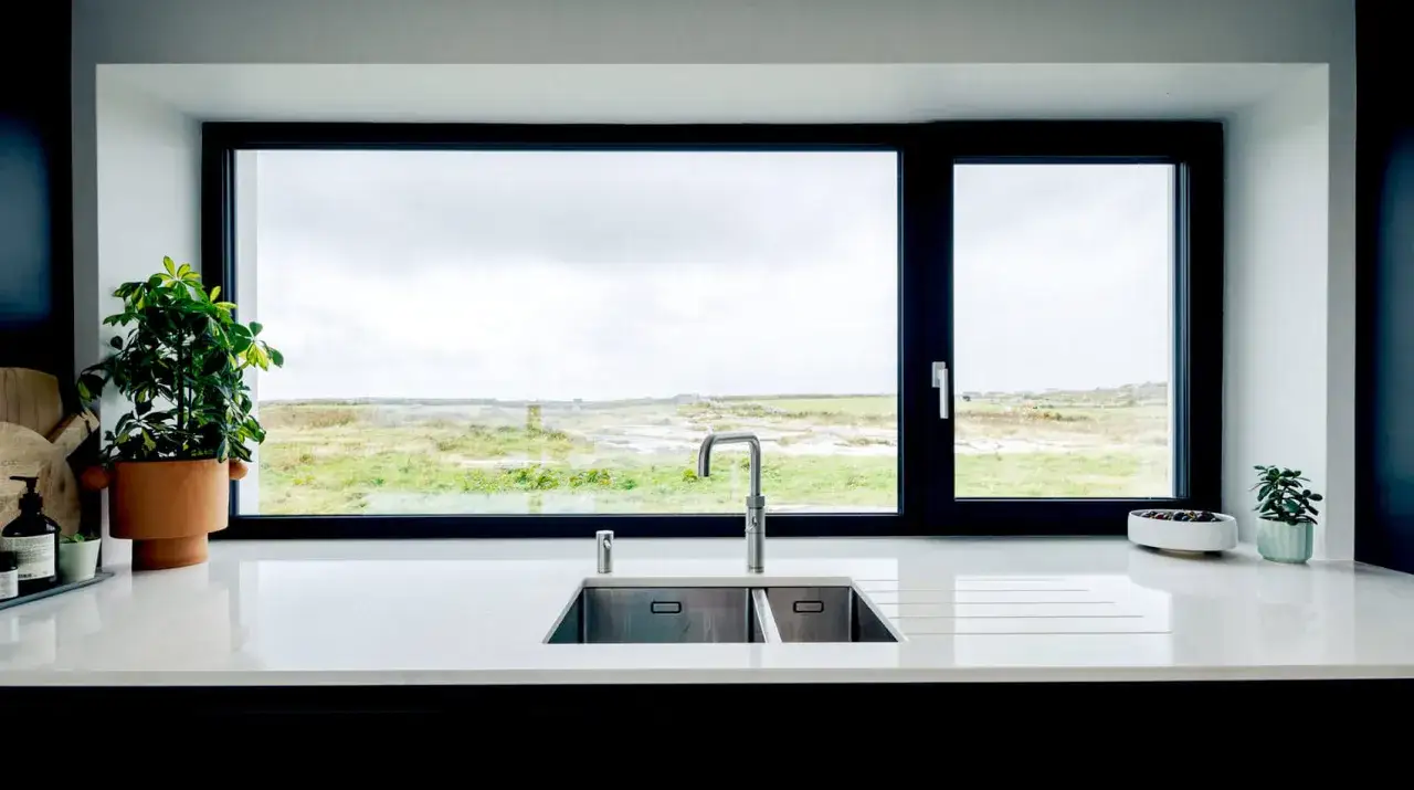Nowoczesna kuchnia z podwójnym zlewozmywakiem i baterią pod dużym oknem, z którego roztacza się widok na zielone pola. Okna w kuchni są na wysokości blatu.