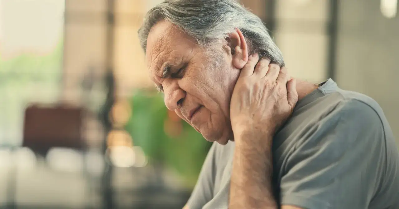 Starszy mężczyzna zamyka oczy, trzymając się za kark. Może to być jeden z objawów zapalenia opon mózgowych.
