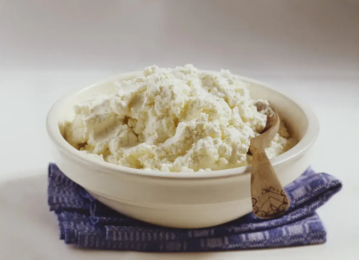 Biały ser w misce z drewnianą łyżką na niebieskiej serwetce.