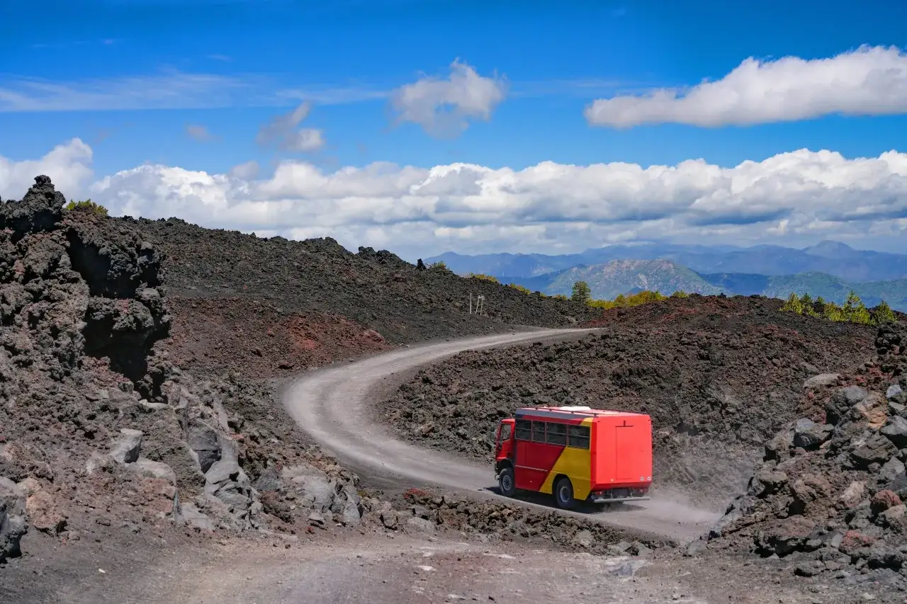 Czerwono-żółty autobus turystyczny jedzie krętą drogą przez wulkaniczne krajobrazy Etny.