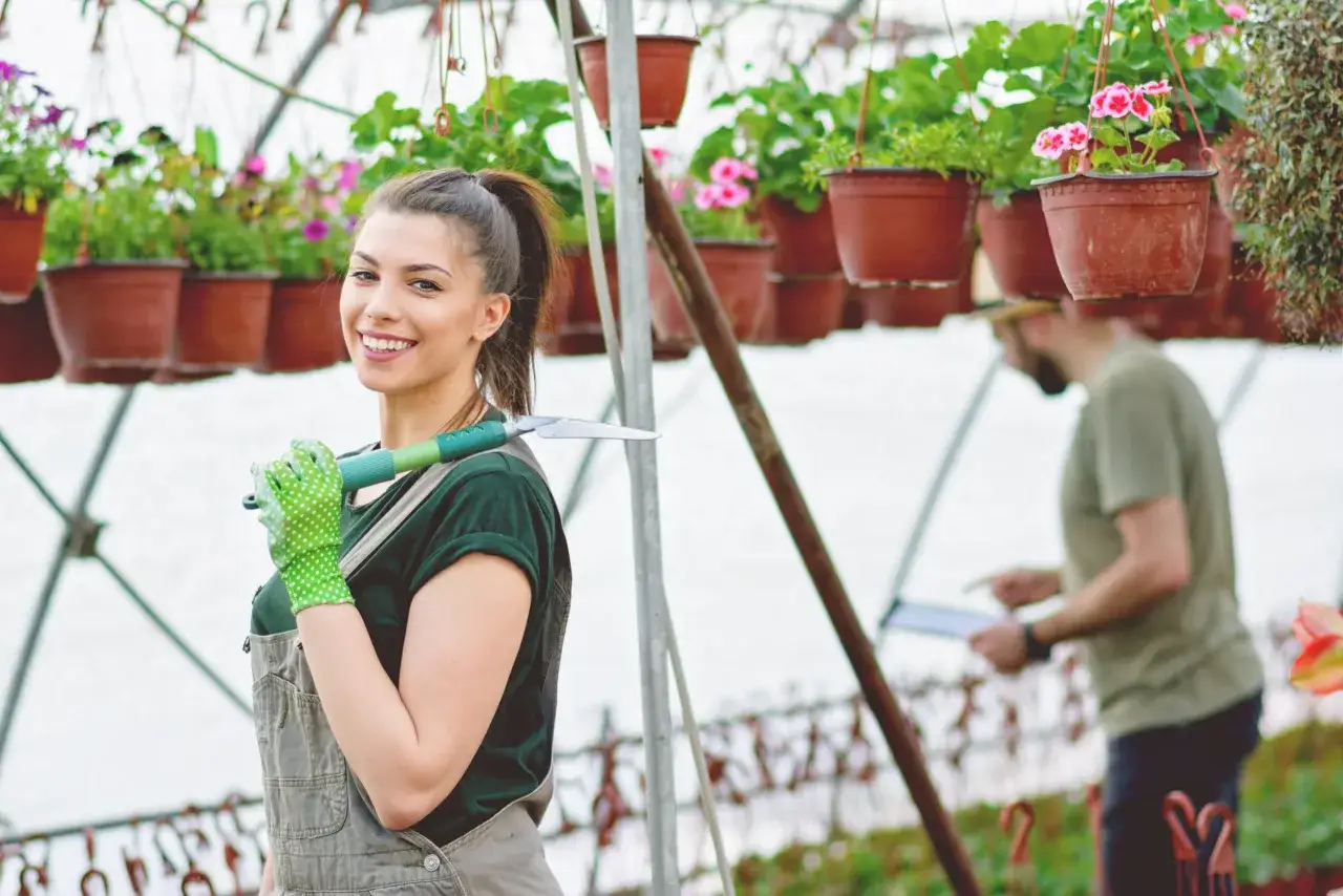 Młoda kobieta w ogrodniczkach i zielonych rękawiczkach z narzędziem w szklarni. Idealne miejsce, gdzie można pracować w wieku 15 lat, to praca przy roślinach.