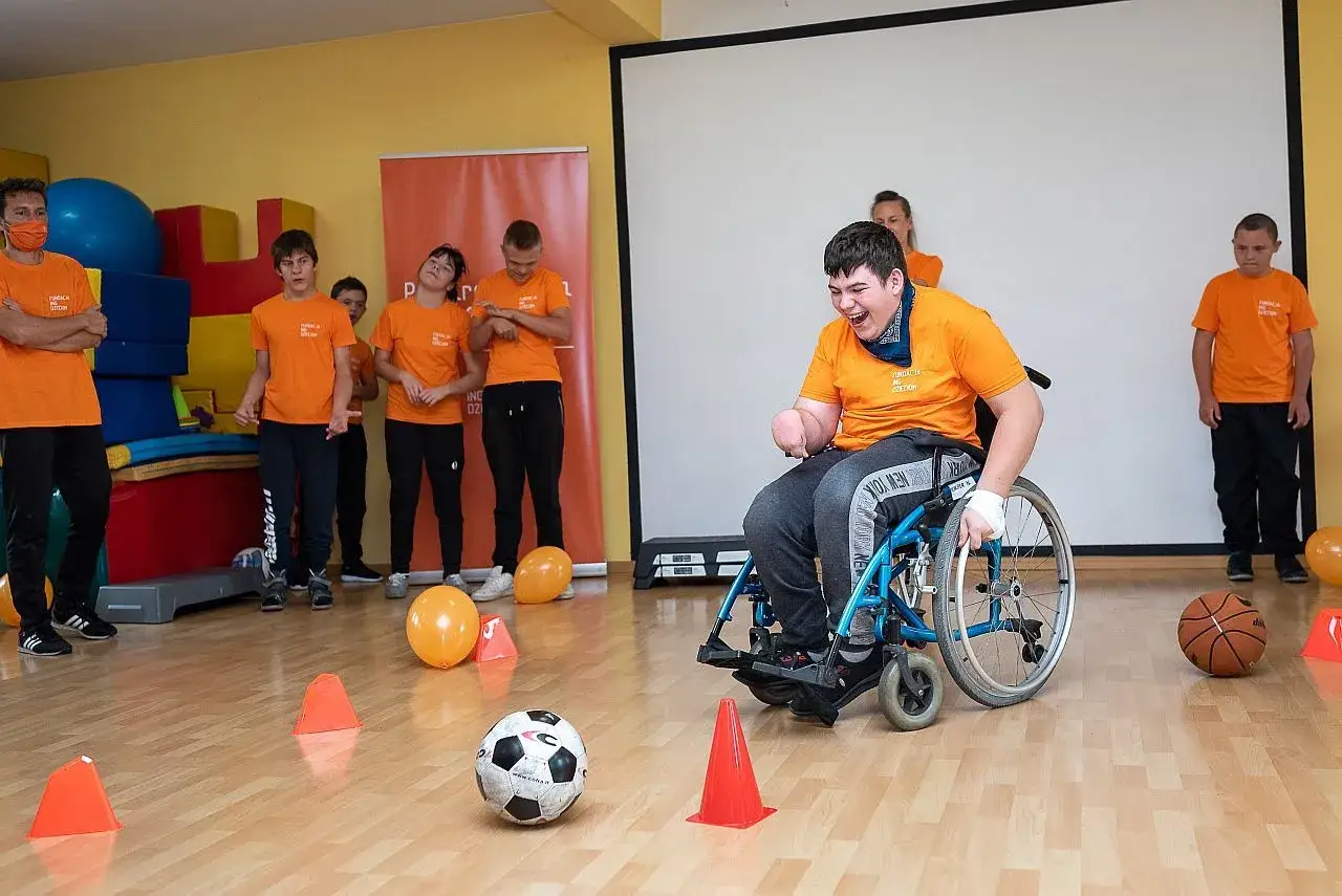 Chłopiec na wózku z uśmiechem gra w piłkę, a obok stoją inni uczestnicy. Widać, że czy na nauczaniu indywidualnym jest WF!