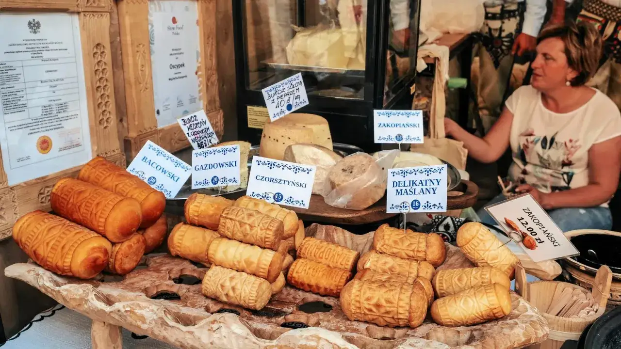 Oscypki i inne sery góralskie na drewnianej desce, z etykietami cenowymi i sprzedawczynią w tle.