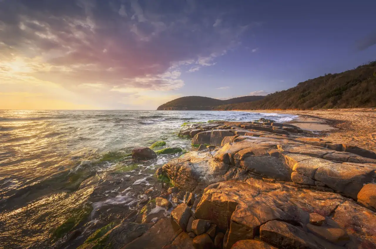Skaliste wybrzeże i morze o zachodzie słońca, z falami rozbijającymi się o kamienie.