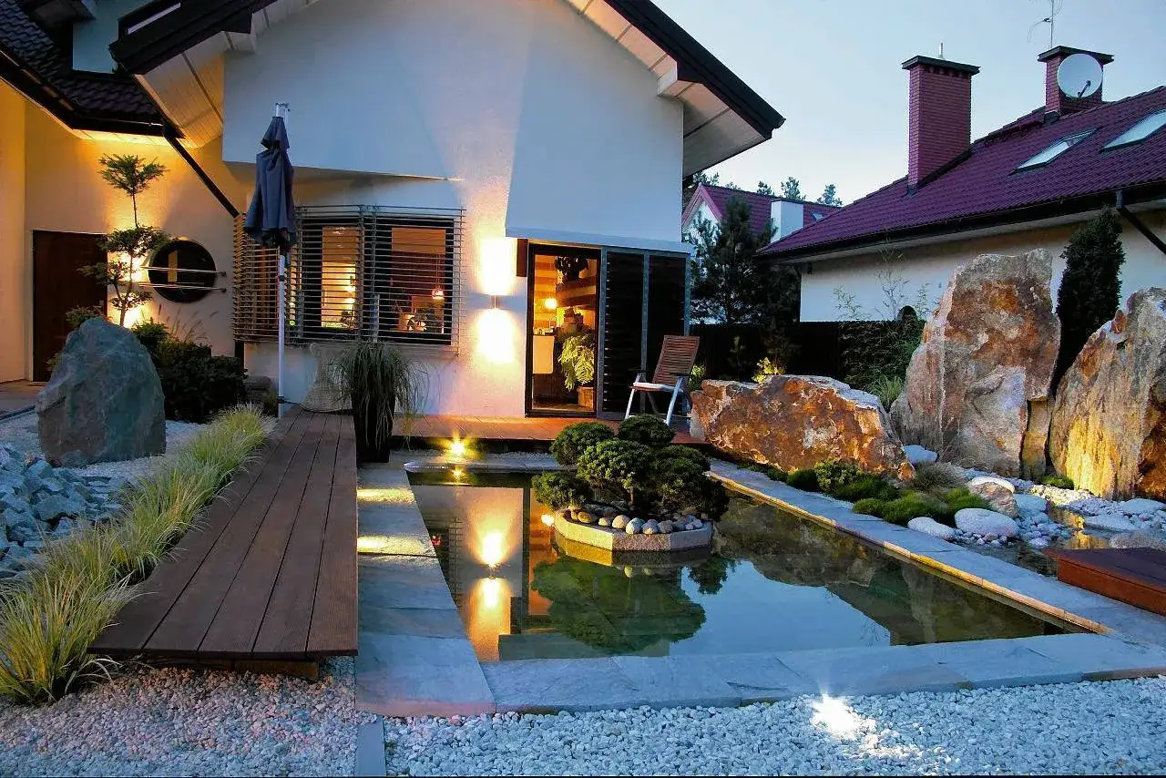 Oświetlenie zewnętrzne domu jednorodzinnego tworzy magiczną atmosferę wokół oczka wodnego i skalnych formacji.