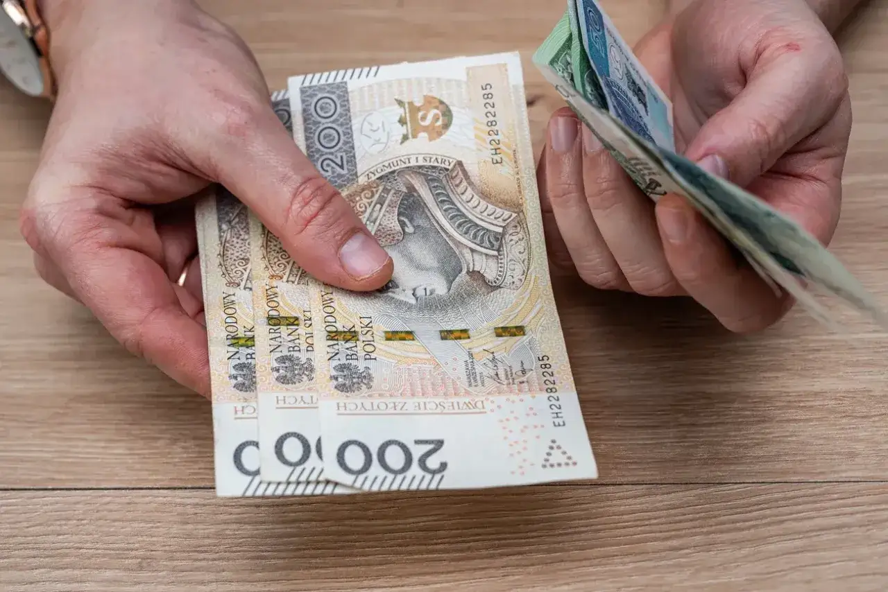 Dłonie liczą polskie banknoty. Czy mieszkaniówka wlicza się do dochodu? Pieniądze na drewnianym blacie.