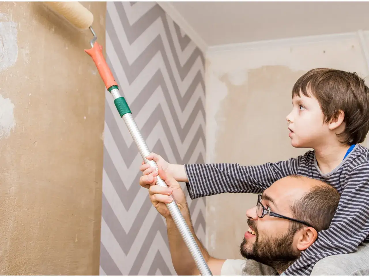 Ojciec i syn wspólnie zajmują się tapetowaniem ścian. Chłopiec pomaga tacie przy wałku.