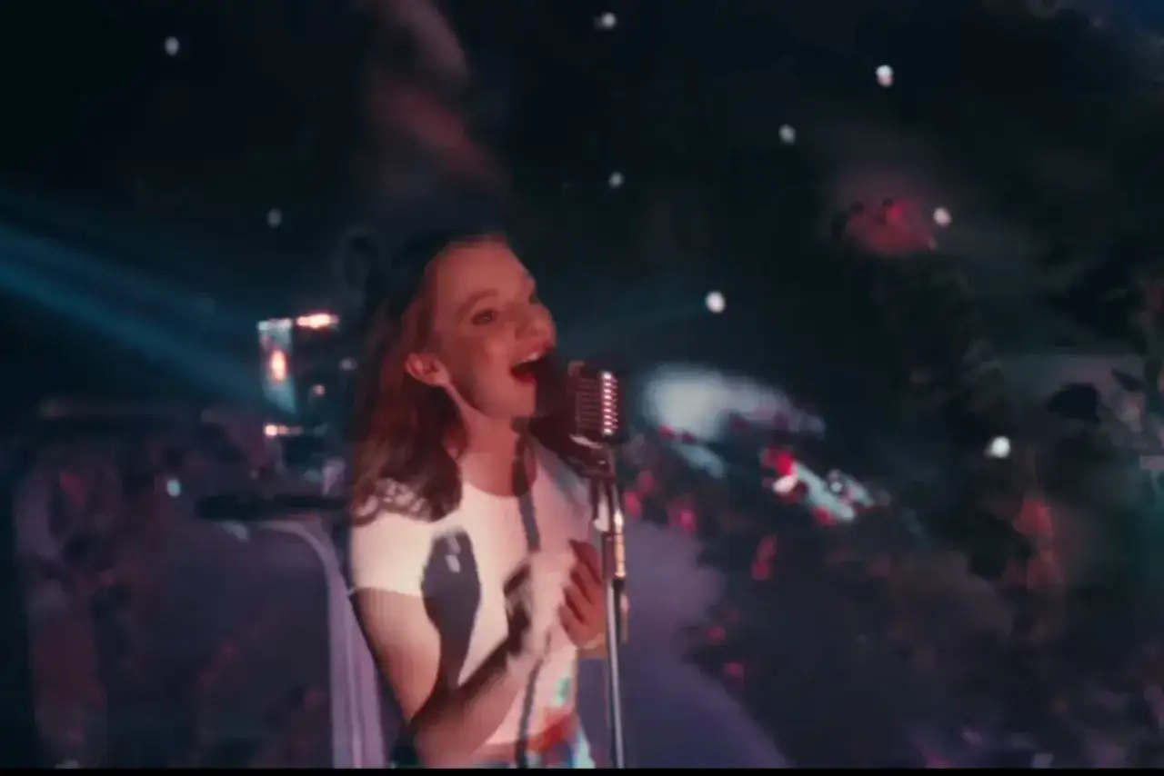 Młoda dziewczyna z rudymi włosami śpiewa do mikrofonu. W tle widać światła i rozmyte kształty, jakby opowiadała, kto śpiewa, jestem twoją bajką.