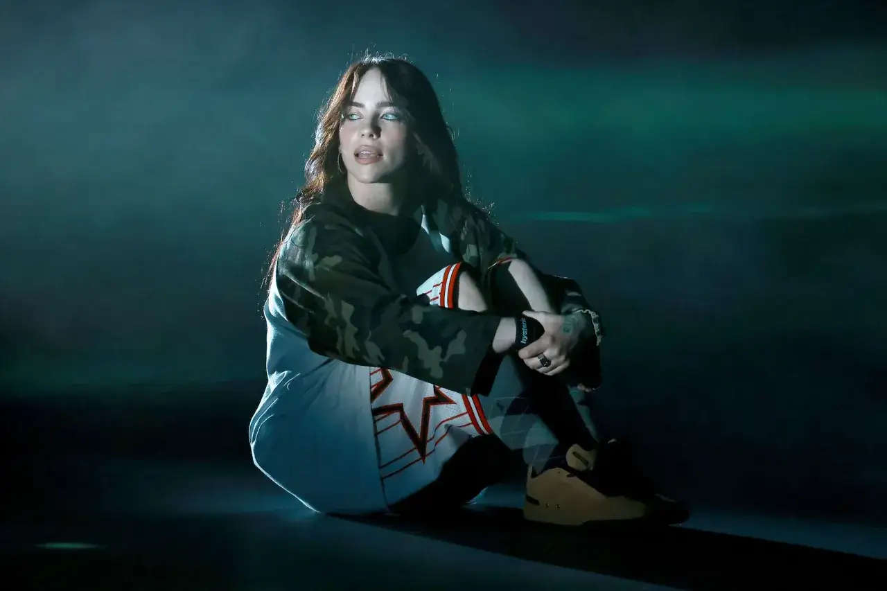 Billie Eilish siedzi na scenie, wpatrzona w dal. Jej stylizacja nawiązuje do estetyki jej albumu.