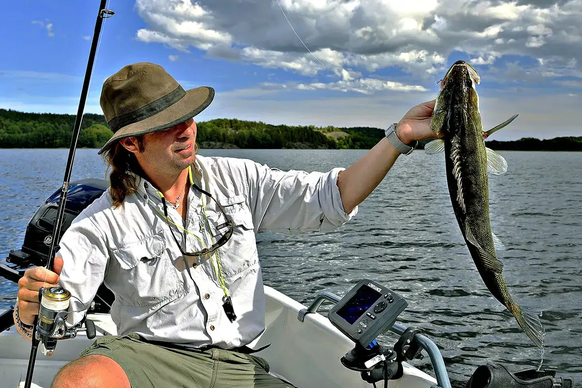 Wędkarz zadowolony z połowu, trzymający rybę na łodzi. Idealna pogoda dla rybaków, błękitne niebo z chmurami.