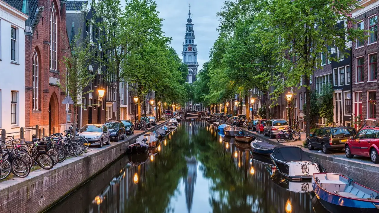 Kanał w Amsterdamie z łodziami, rowerami i wieżą kościelną w tle.