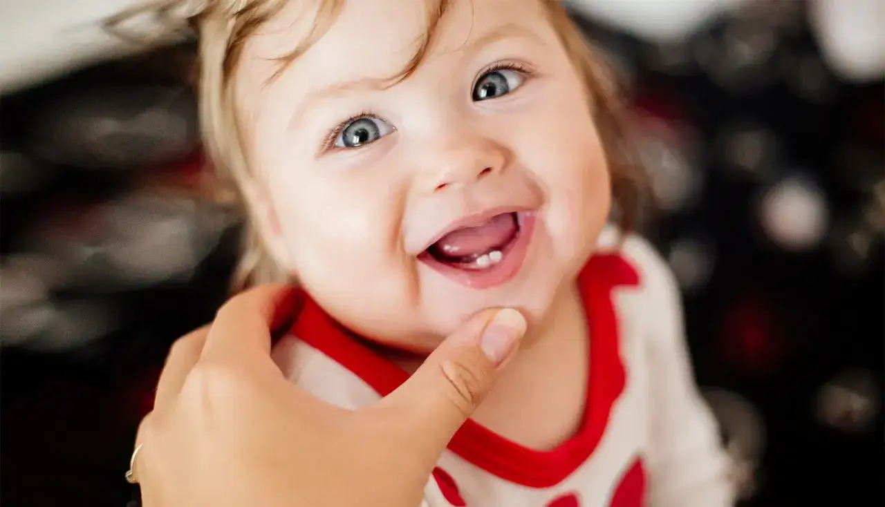 Uśmiechnięte dziecko z widocznymi ząbkami, które właśnie wychodzą. Delikatne dziąsła przy ząbkowaniu są lekko zaczerwienione.