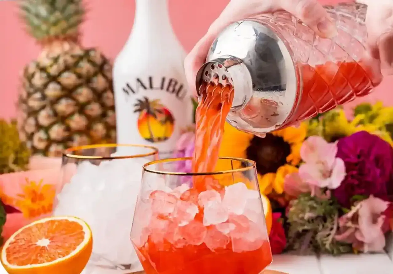 Malibu z sokiem ananasowym nalewany do szklanki z lodem. W tle ananas, butelka Malibu i kwiaty.