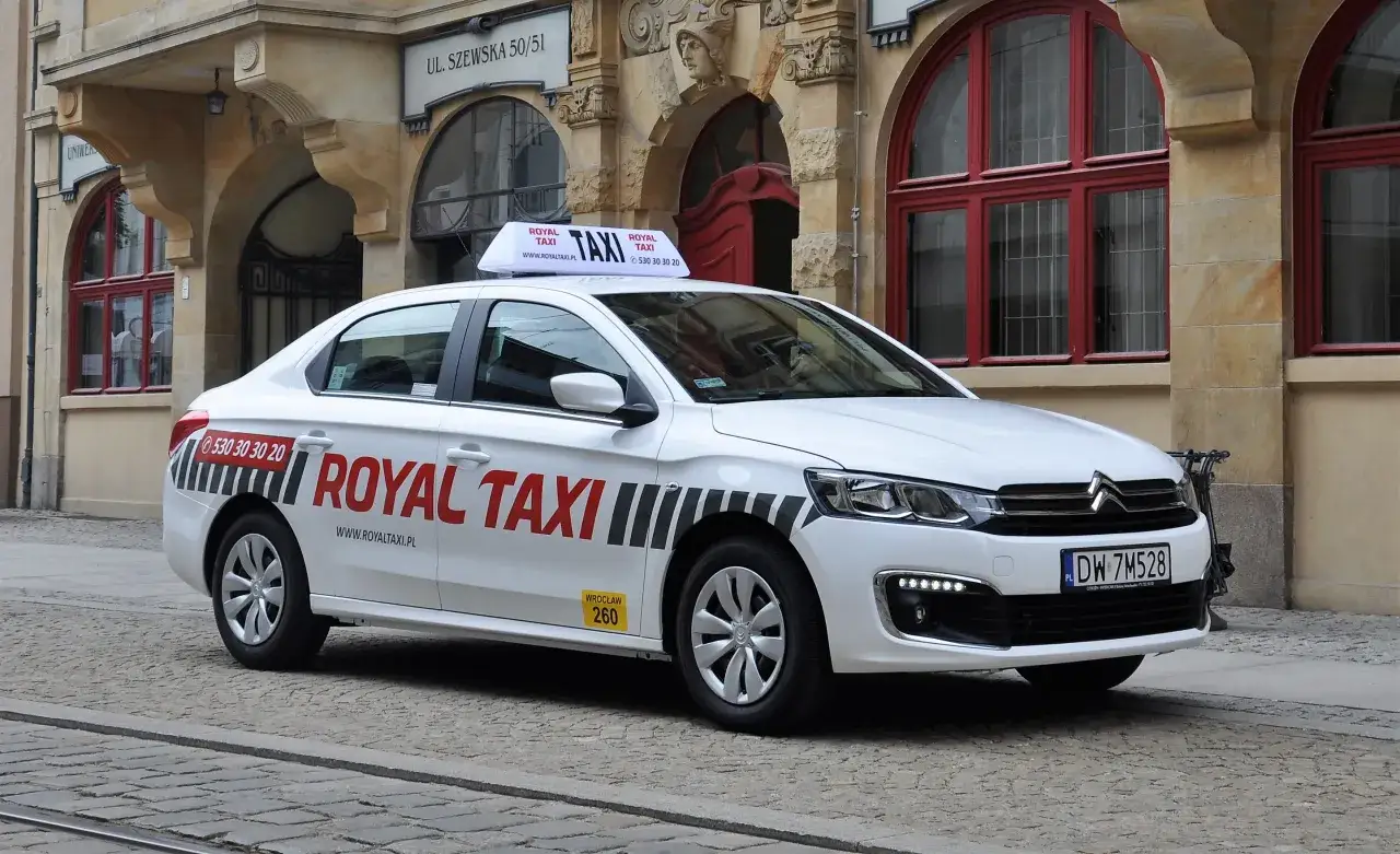 Biała taksówka Royal Taxi z numerem 260 na tle zabytkowej kamienicy we Wrocławiu.