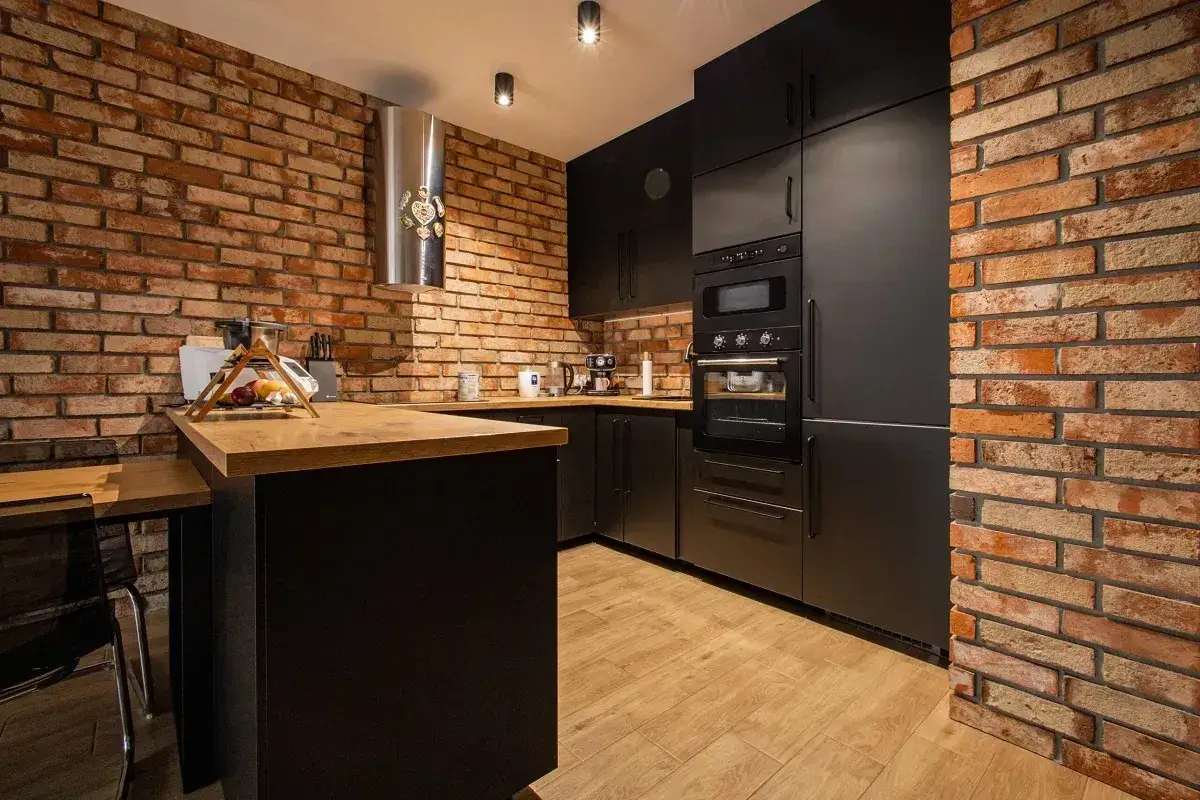 Nowoczesna kuchnia z czarnymi meblami i blatem z litego drewna, kontrastująca z rustykalną ścianą z cegły.