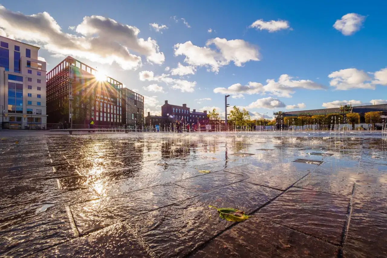 Zachód słońca nad miejską fontanną, odbijający się w mokrej nawierzchni. Czułość filmu uchwycona w złotych promieniach i tańczących kroplach wody.
