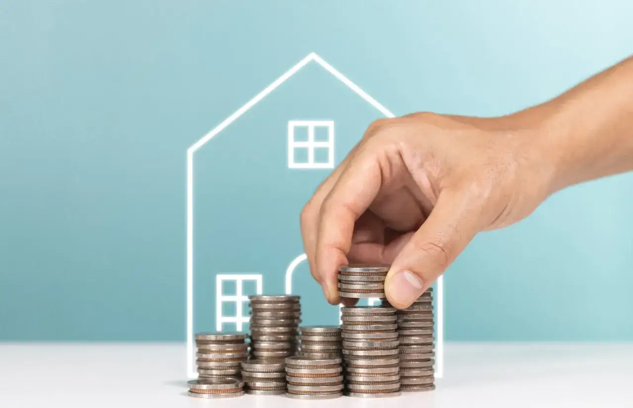 Dłoń układa stos monet na tle zarysu domu.