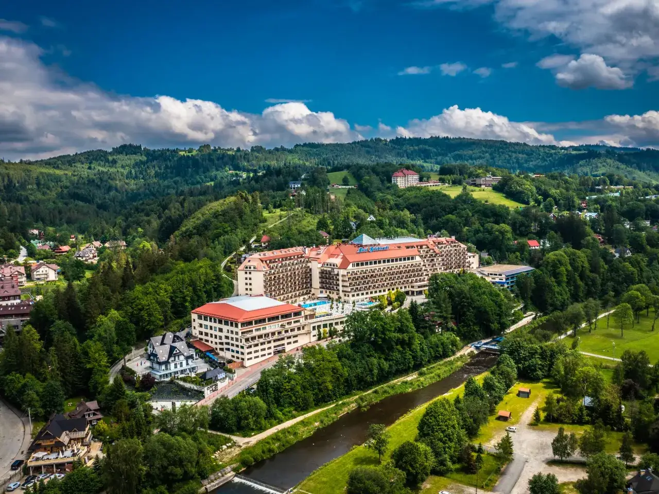Widok z lotu ptaka na duży hotel Gołębiewski otoczony zielonymi wzgórzami i rzeką.