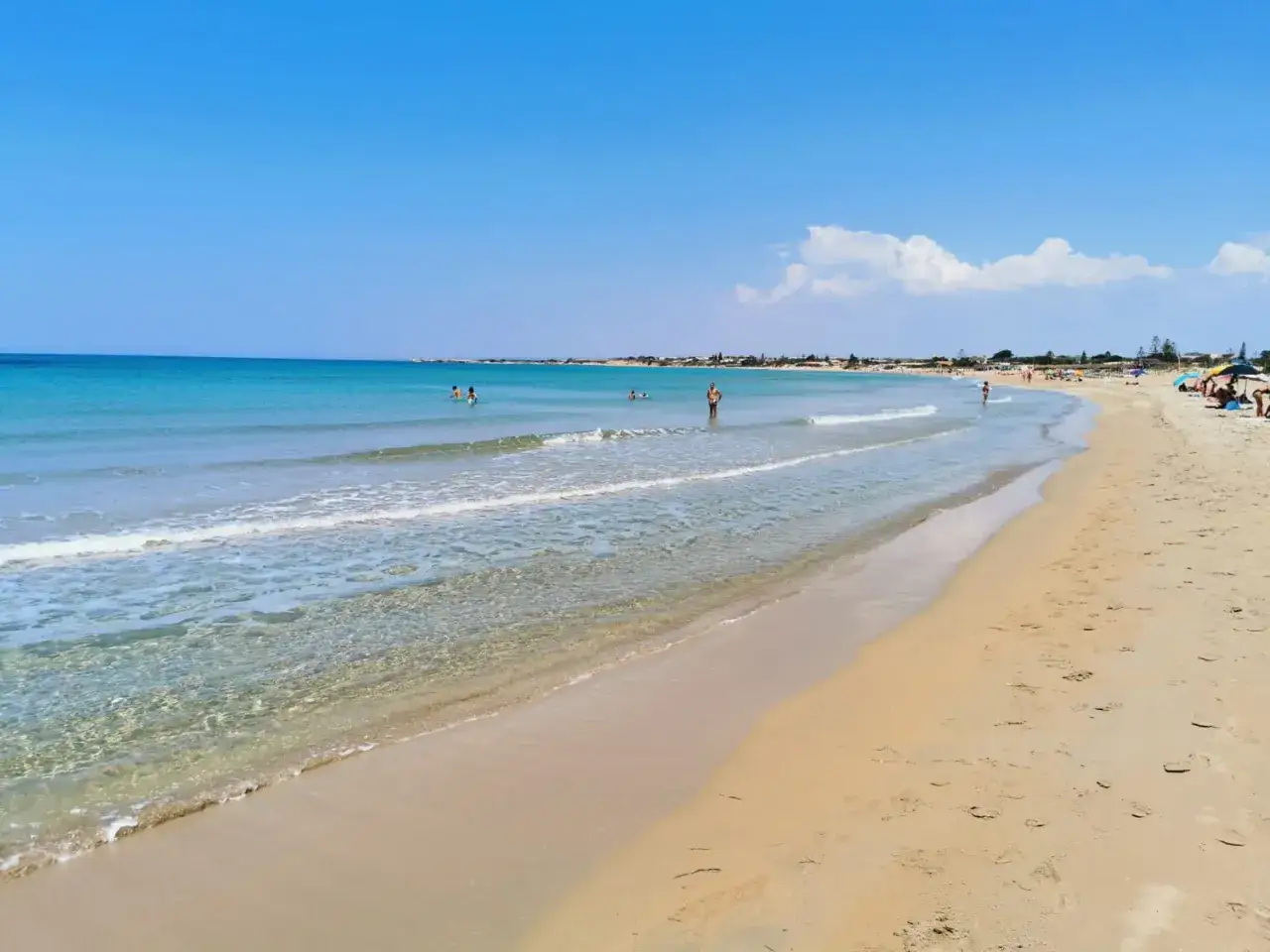 A wide, sandy beach with clear turquoise water and a few people swimming and sunbathing under a bright blue sky.