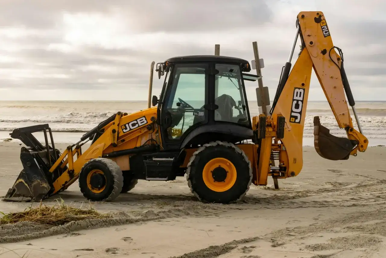 Żółta koparko-ładowarka firmy JCB stoi na piaszczystej plaży, gotowa do pracy.