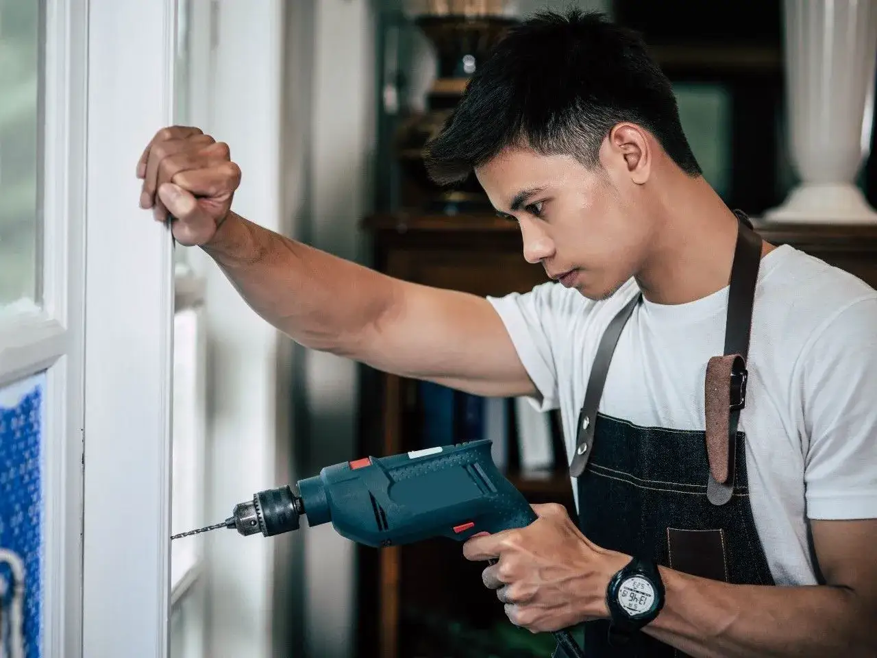 Mężczyzna w fartuchu wierci w białej ramie drzwi wiertarką.