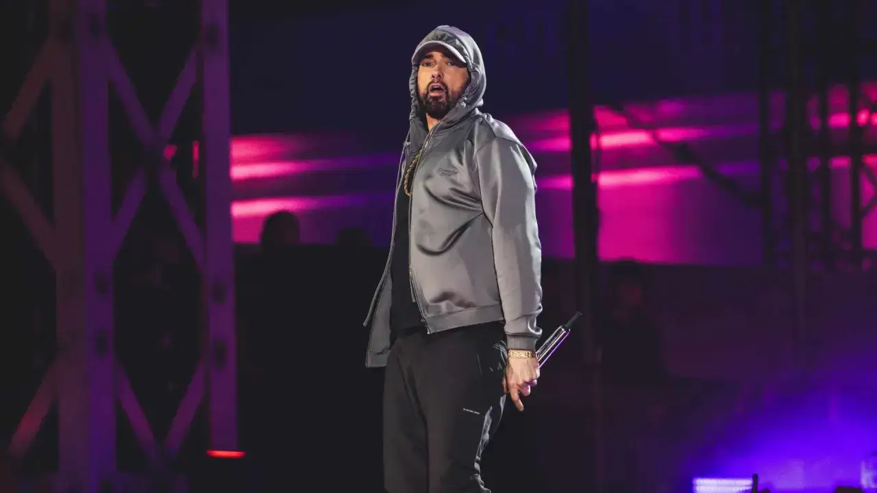 Eminem na scenie w szarej bluzie z kapturem i czarnych spodniach, trzymający mikrofon.
