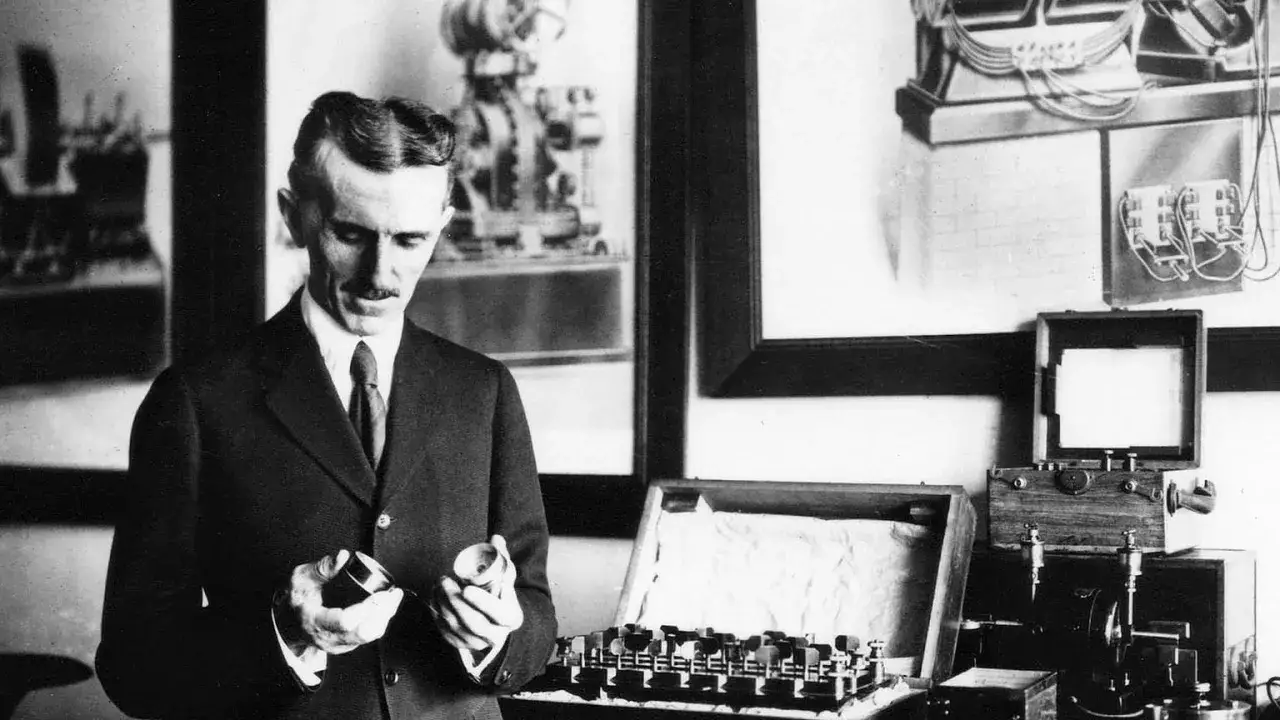 Nikola Tesla w garniturze trzymający elementy aparatury elektrycznej, obok skrzynki z urządzeniami.