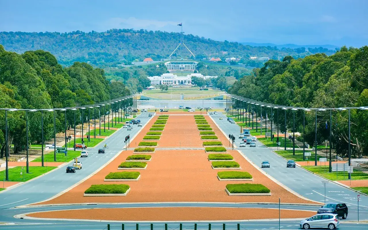 Widok na Parliament House, symbol tego, co jest stolicą Australii. Długa aleja z drzewami i samochodami prowadzi do budynku.