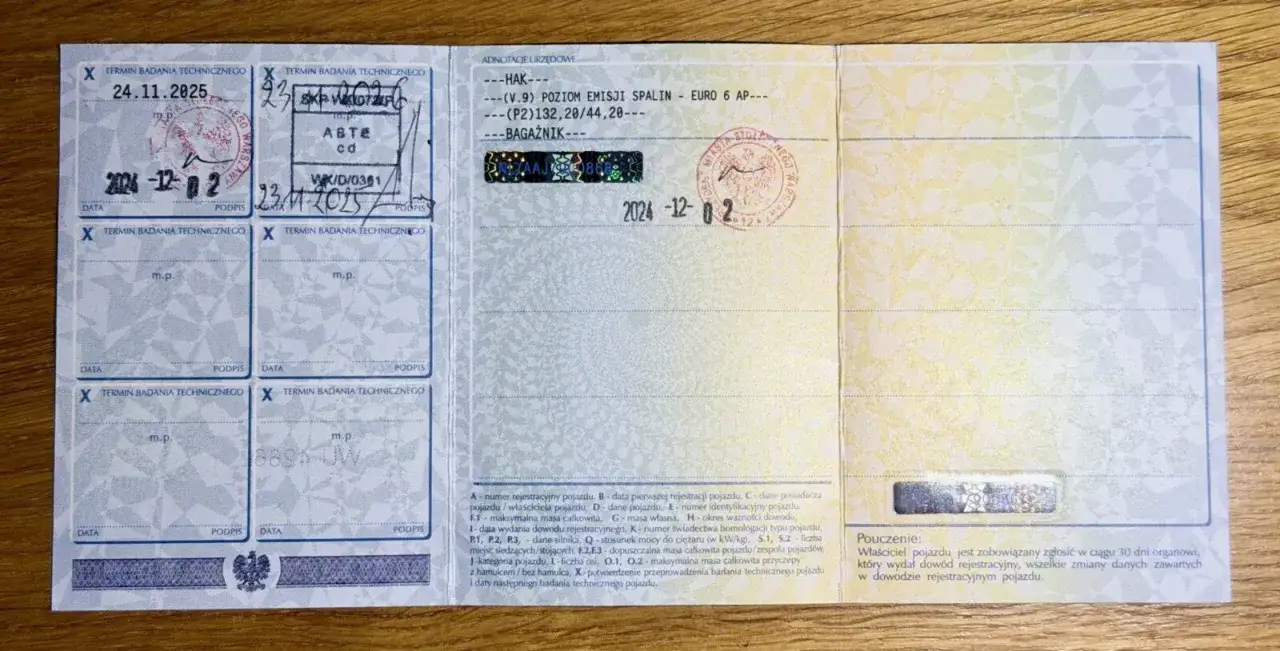Fragment dowodu rejestracyjnego pojazdu z pieczątkami i wpisami, widoczny na drewnianym tle.