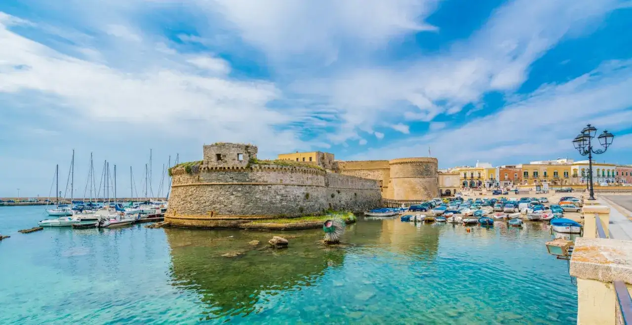 A stone castle overlooks a harbor filled with boats and yachts under a bright blue sky.