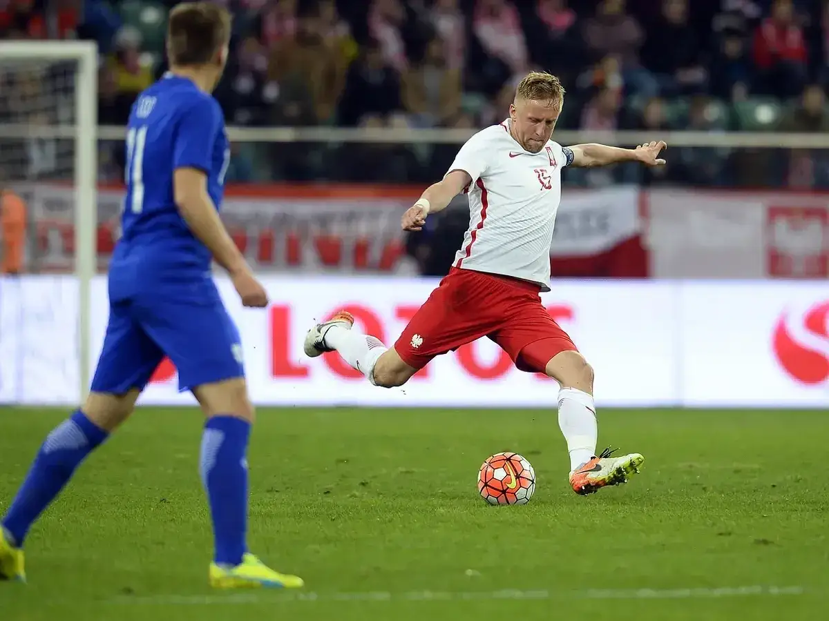 Piłkarz w białej koszulce i czerwonych spodenkach kopie piłkę na boisku.