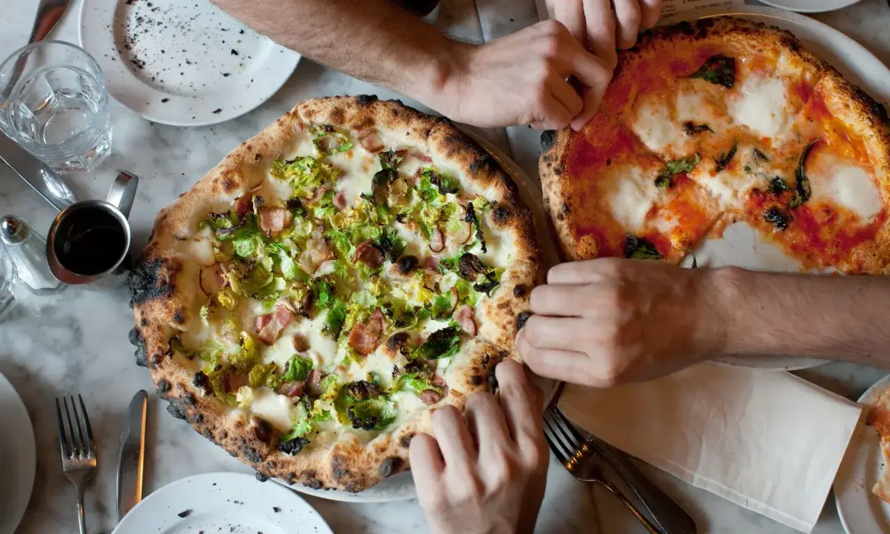 Dwie pizze na stole, jedna z brukselką i boczkiem, druga margherita. Ludzie sięgają po kawałki, pokazując, ile wyrasta ciasto na pizze.