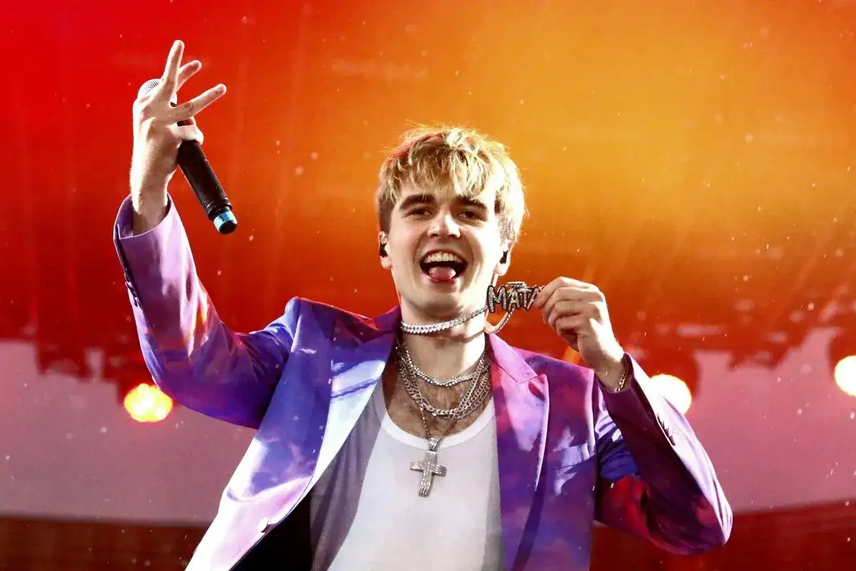 Mata w fioletowej marynarce z naszyjnikiem z napisem "MATA" i mikrofonem na koncercie.