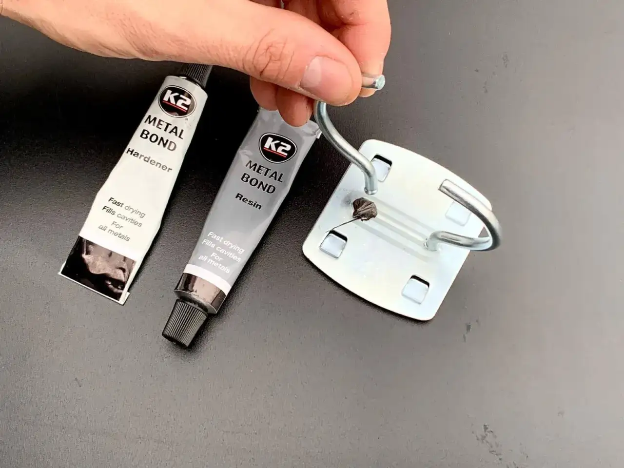 Dwie tubki kleju K2 Metal Bond (utwardzacz i żywica) oraz metalowy element z zaczepem.