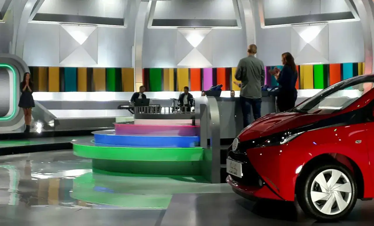 Studio telewizyjne, gdzie nagrywane jest koło fortuny. Widać scenografię z kolorowymi panelami, schodami i czerwonym samochodem.