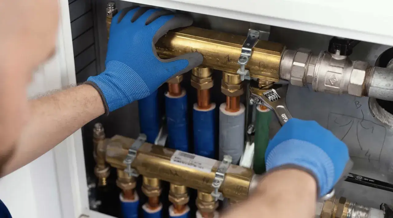 Ręce w niebieskich rękawiczkach montują instalację hydrauliczną, używając klucza.