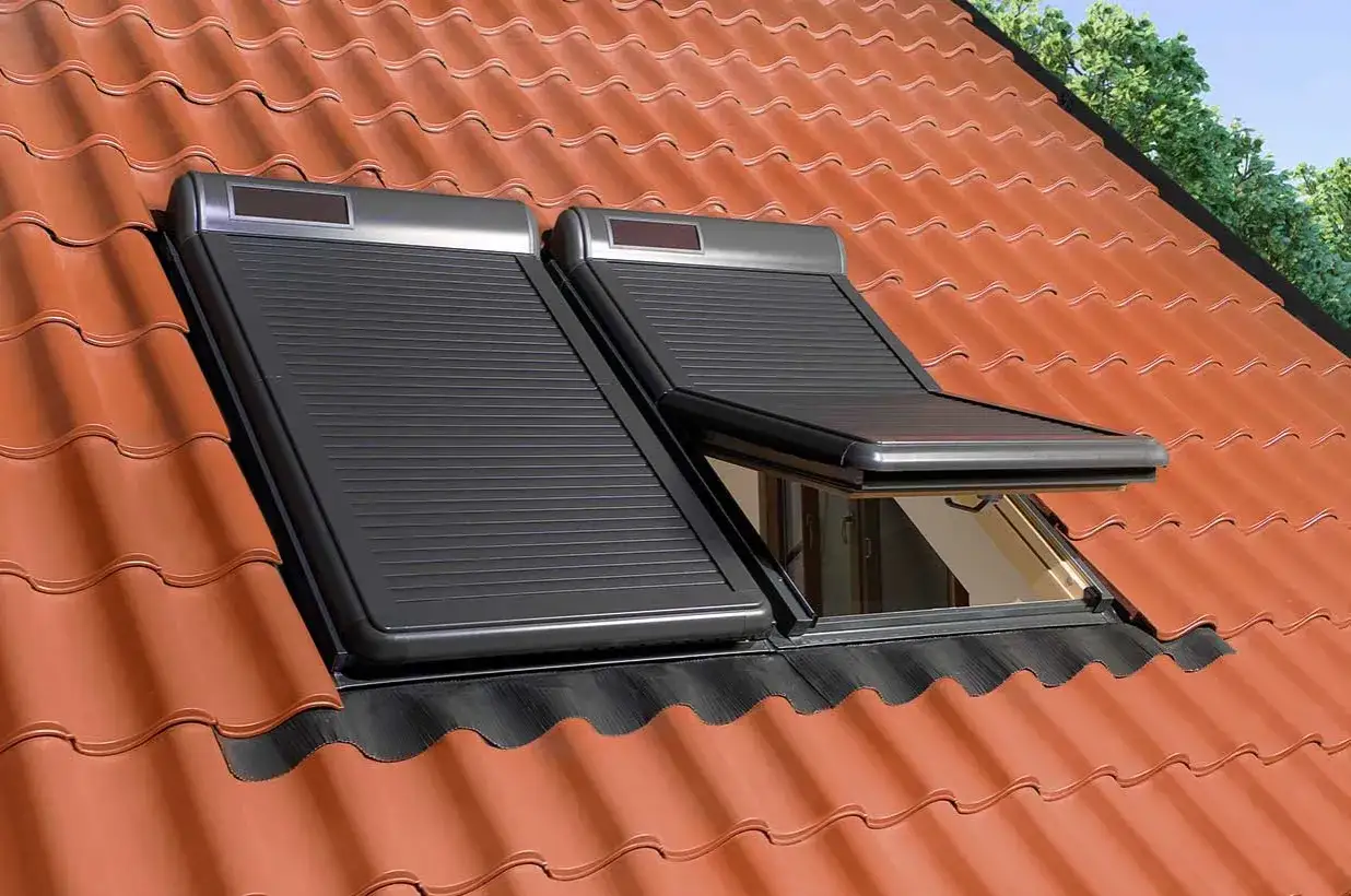 Dwa okna dachowe z czarną roletą zewnętrzną na dachu pokrytym dachówką. Jedna roleta jest częściowo opuszczona.