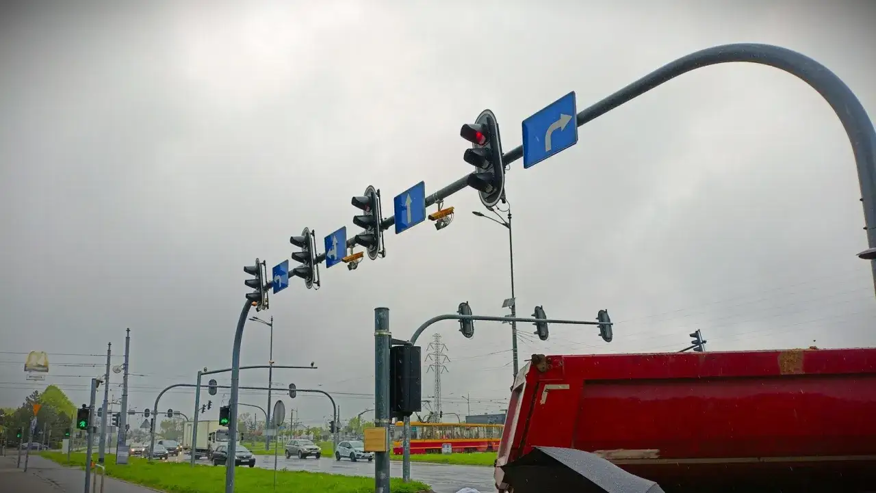 Na skrzyżowaniu wiszą sygnalizatory świetlne i znaki kierunkowe. Widać żółty tramwaj i czerwony wywrotkę. Gdzie są kamery rejestrujące przejazd na czerwonym świetle?