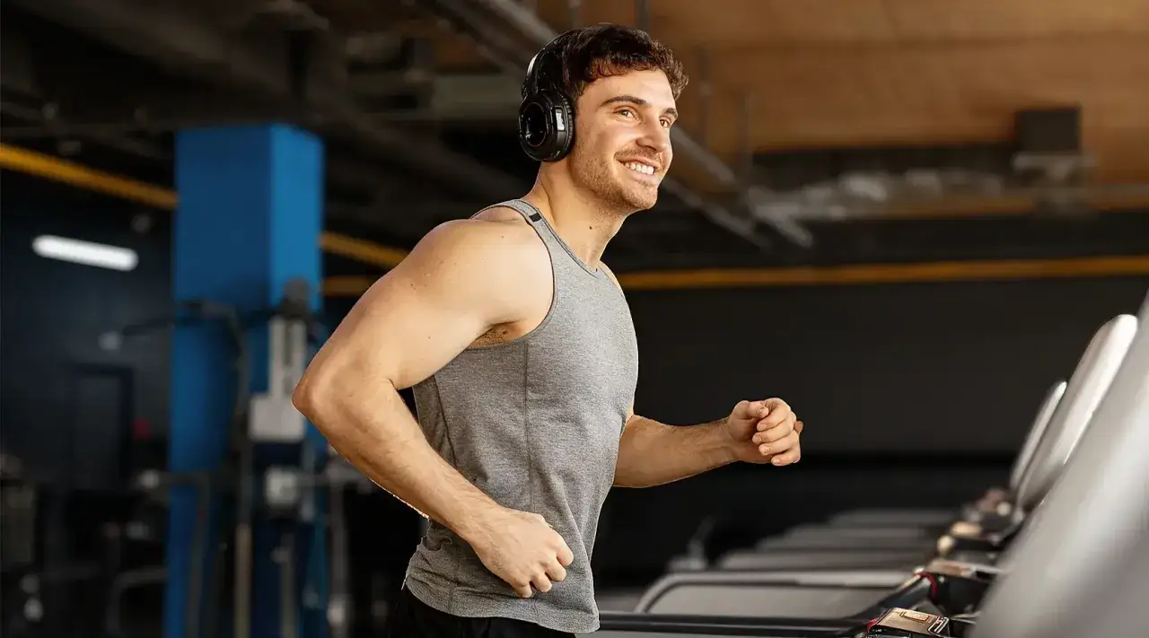 Uśmiechnięty mężczyzna w słuchawkach biegnie na bieżni w siłowni.