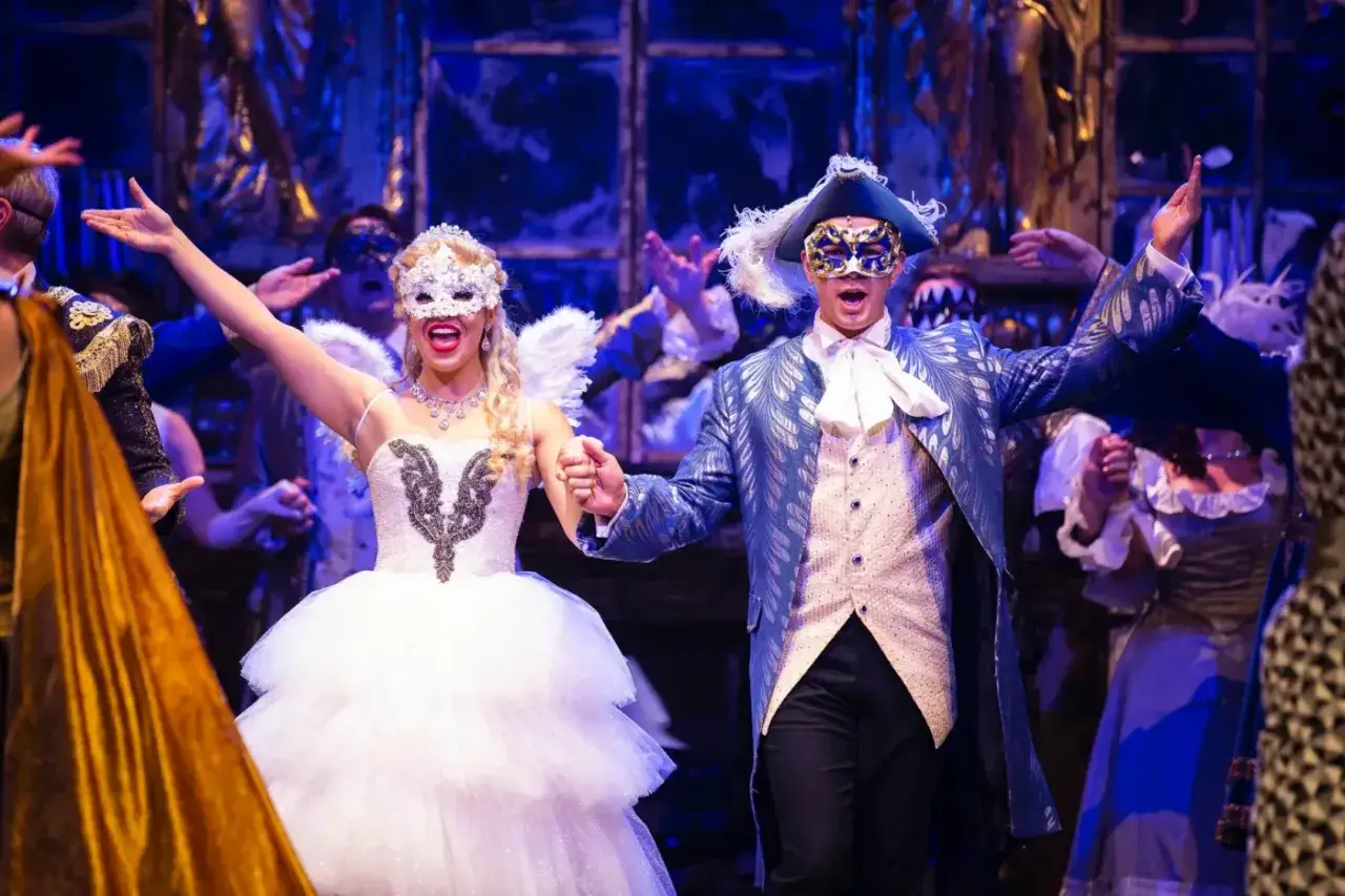 Upiór w Operze Warszawa: para w maskach i strojach z epoki tańczy na balu. Ona w białej sukni i z anielskimi skrzydłami, on w błękitnym fraku.