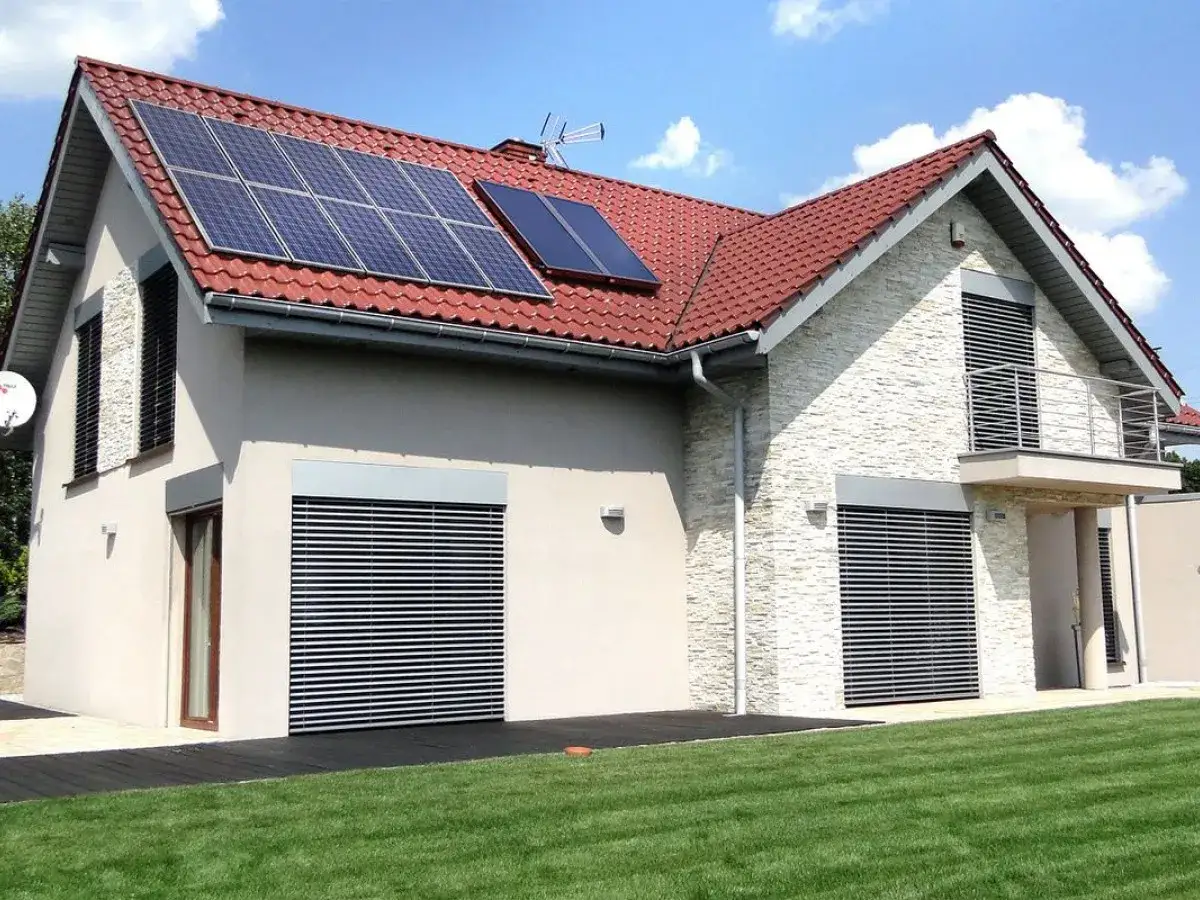 Dom z panelami fotowoltaicznymi na czerwonym dachu, z zielonym trawnikiem przed budynkiem.