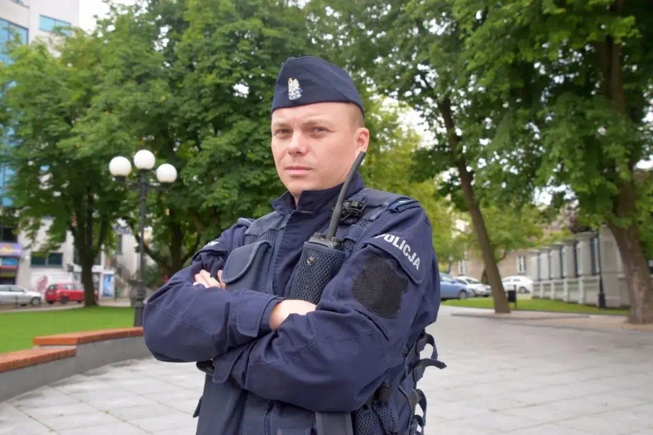Policjant w granatowym mundurze, w sprawie umundurowania policjantów, stoi na tle zieleni.
