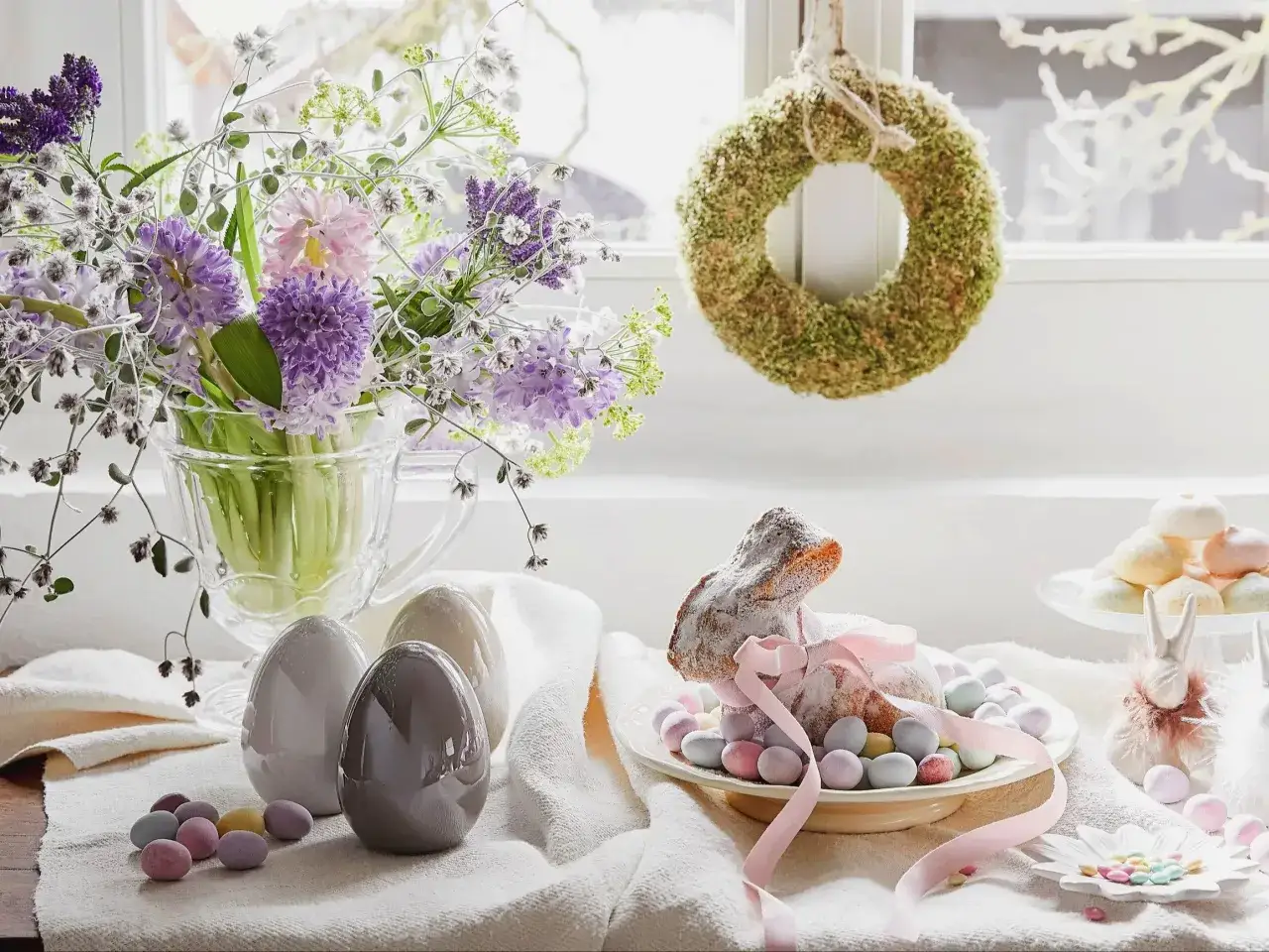 Wielkanocne dekoracje: bukiet wiosennych kwiatów, ceramiczne jajka, lukrowany zajączek otoczony cukierkami i mechowy wianek.