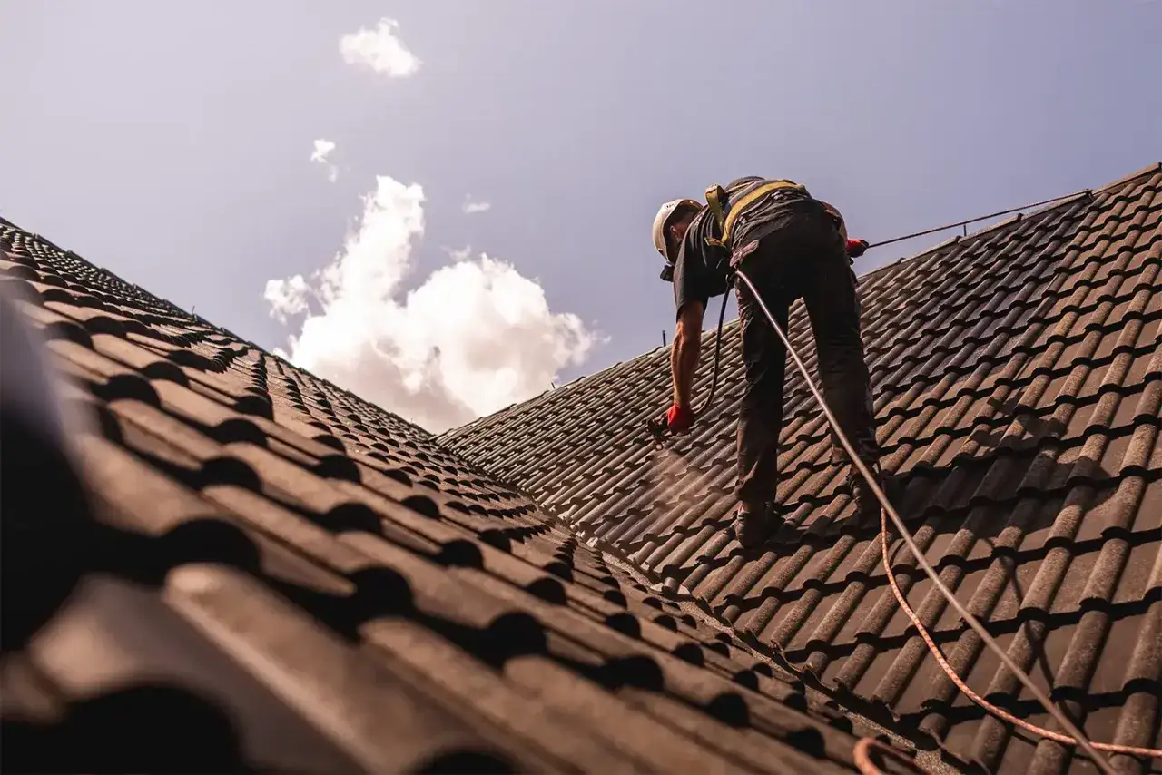 Pracownik w kasku i uprzęży zabezpieczającej wykonuje renowację dachu, natryskując dachówkę.