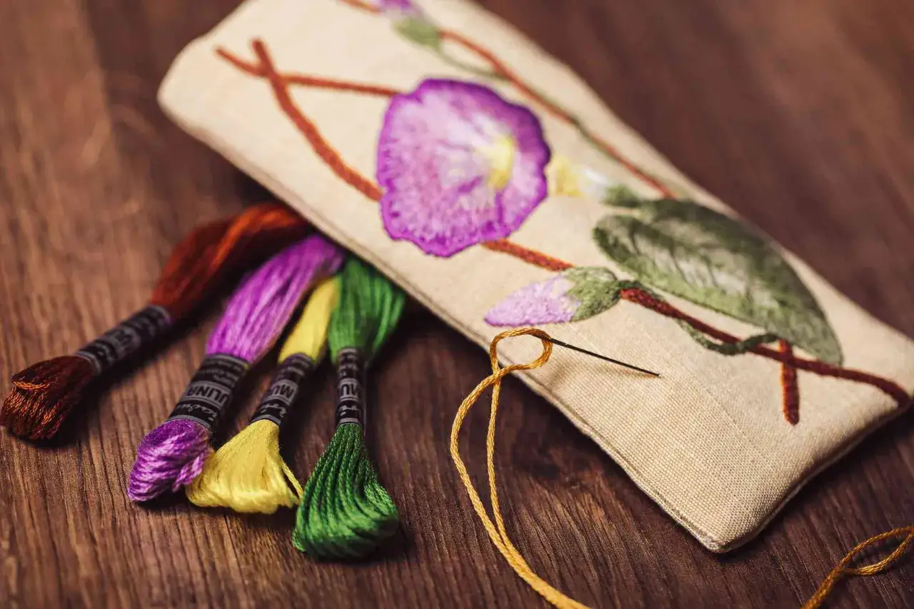 Kolorowe nici do haftowania obok haftowanego kwiatu na płótnie. Igła z żółtą nitką w ręku.