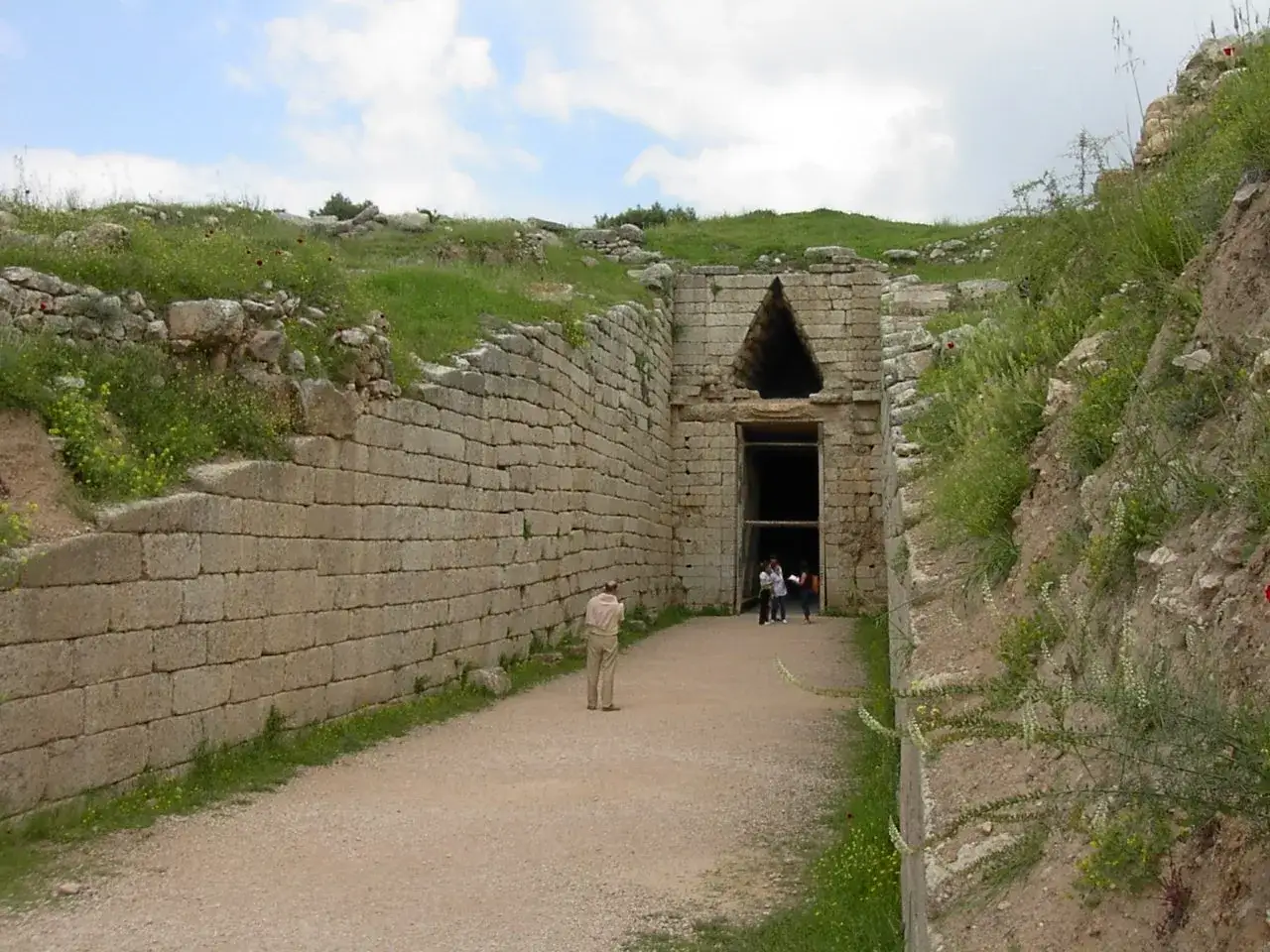 Kamienna ściana prowadzi do wejścia do grobowca Agamemnona. W oddali widać ludzi wchodzących do środka.
