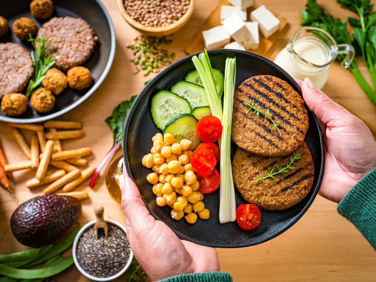 Talerz z wegańskimi burgerami, ciecierzycą, pomidorkami i ogórkiem. W tle inne wegańskie produkty.