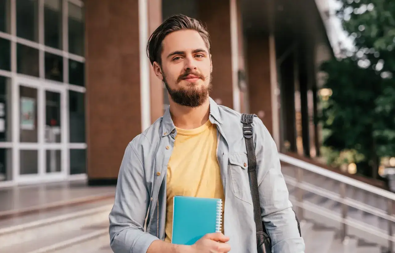 Młody mężczyzna z zeszytem przed budynkiem uczelni. Czy szkoła policealna daje status studenta?