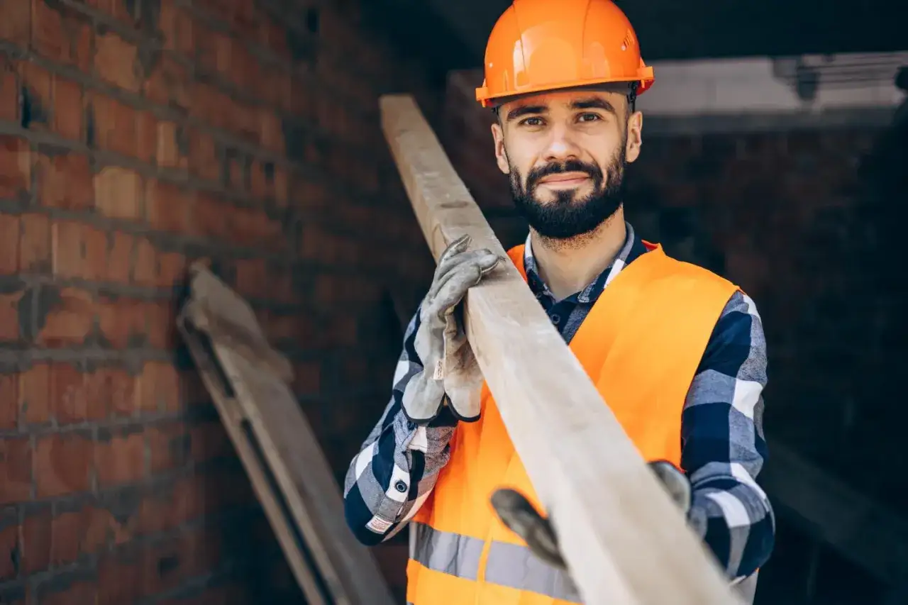 Budowlaniec w kasku i kamizelce niesie drewnianą belkę, przygotowując się do montażu do elewacji.