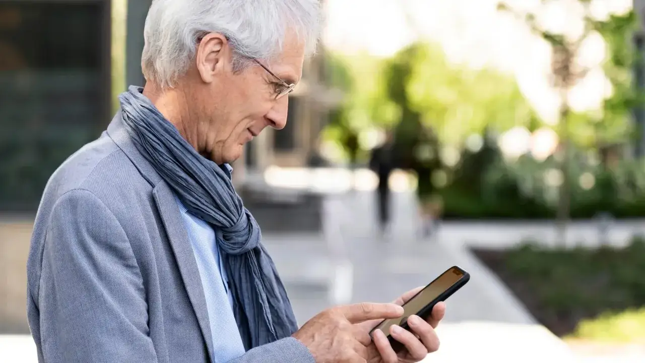 Starszy mężczyzna w okularach i szaliku korzysta ze smartfona, ucząc się, jak połączyć aparat słuchowy z telefonem.